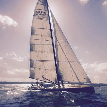 Ein elegantes Segelboot mit großen weißen Segeln von den Online-Segelmachern von Sails24 fährt bei sonnigem Wetter über das offene Meer. Die Sonne scheint direkt hinter dem Mast durch die Segel.