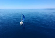 Ein Segelboot fotografiert aus der Vogelperspektive mit einem blauen Spinnaker, von Sailselct, das allein auf einer ruhigen Meeresoberfläche unter klarem Himmel segelt.